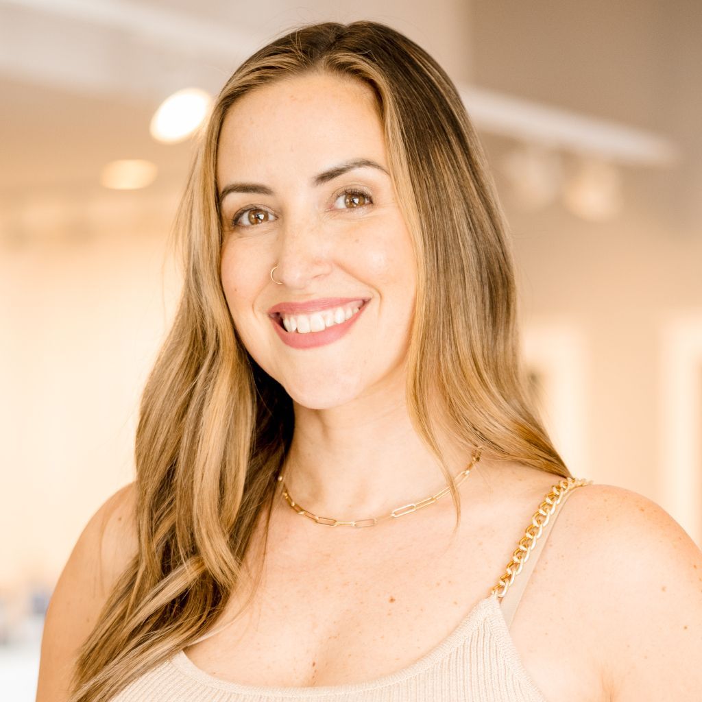 Smiling woman with long hair in a beige top, standing indoors in a softly lit room.