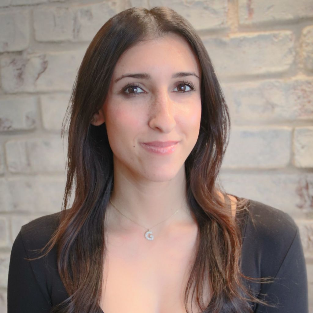 Portrait of a smiling woman with long dark hair, wearing a black top, against a light brick wall.