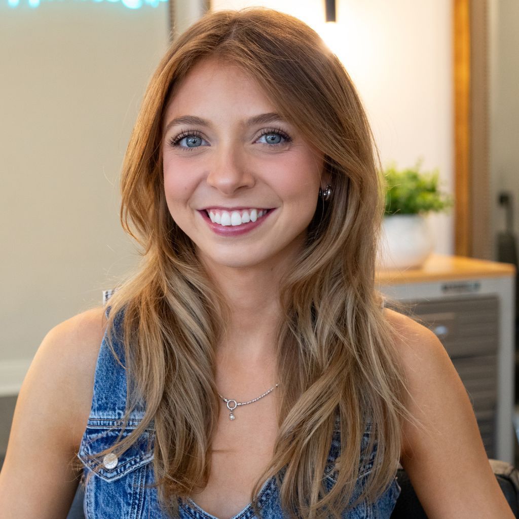Smiling woman in a denim top with long blonde hair, seated indoors with a softly lit background