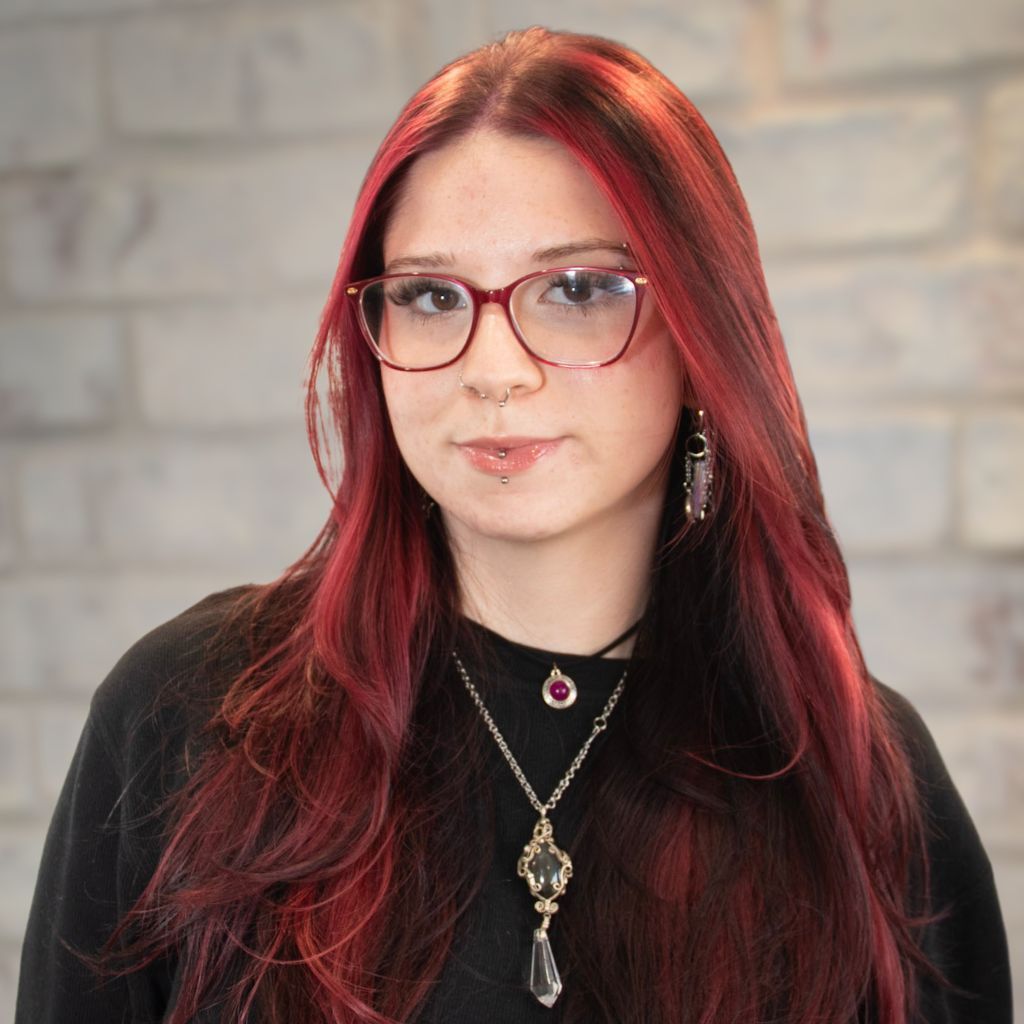 Portrait of a person with red hair, glasses, and layered necklaces against a stone wall backdrop