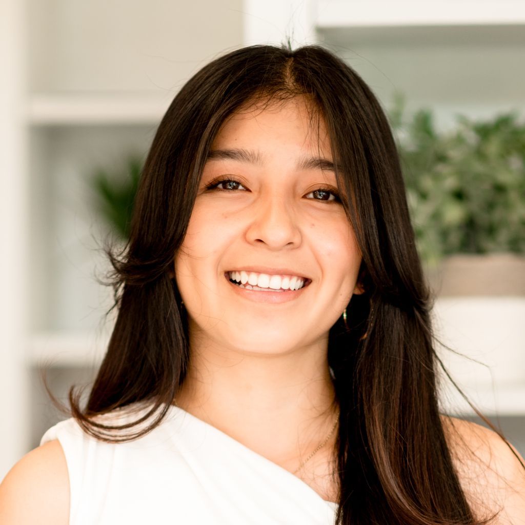 Smiling woman in a white top, with wavy dark hair, seated indoors with plants in the background