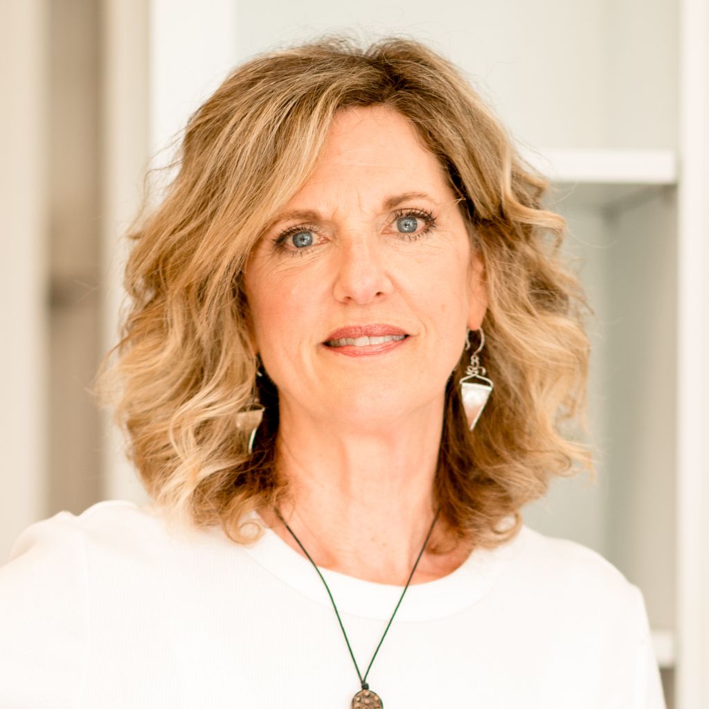 Portrait of a woman with wavy blonde hair wearing a white top and necklace against a bright indoor background