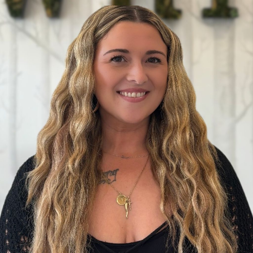 Smiling woman with wavy blonde hair wearing a black top and gold necklace, standing indoors.