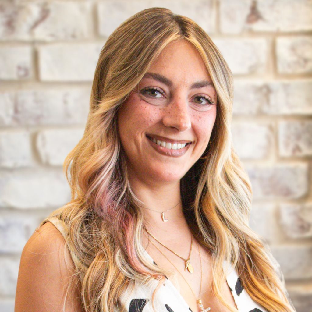 Smiling woman with long wavy blonde hair in a white top against a light brick wall