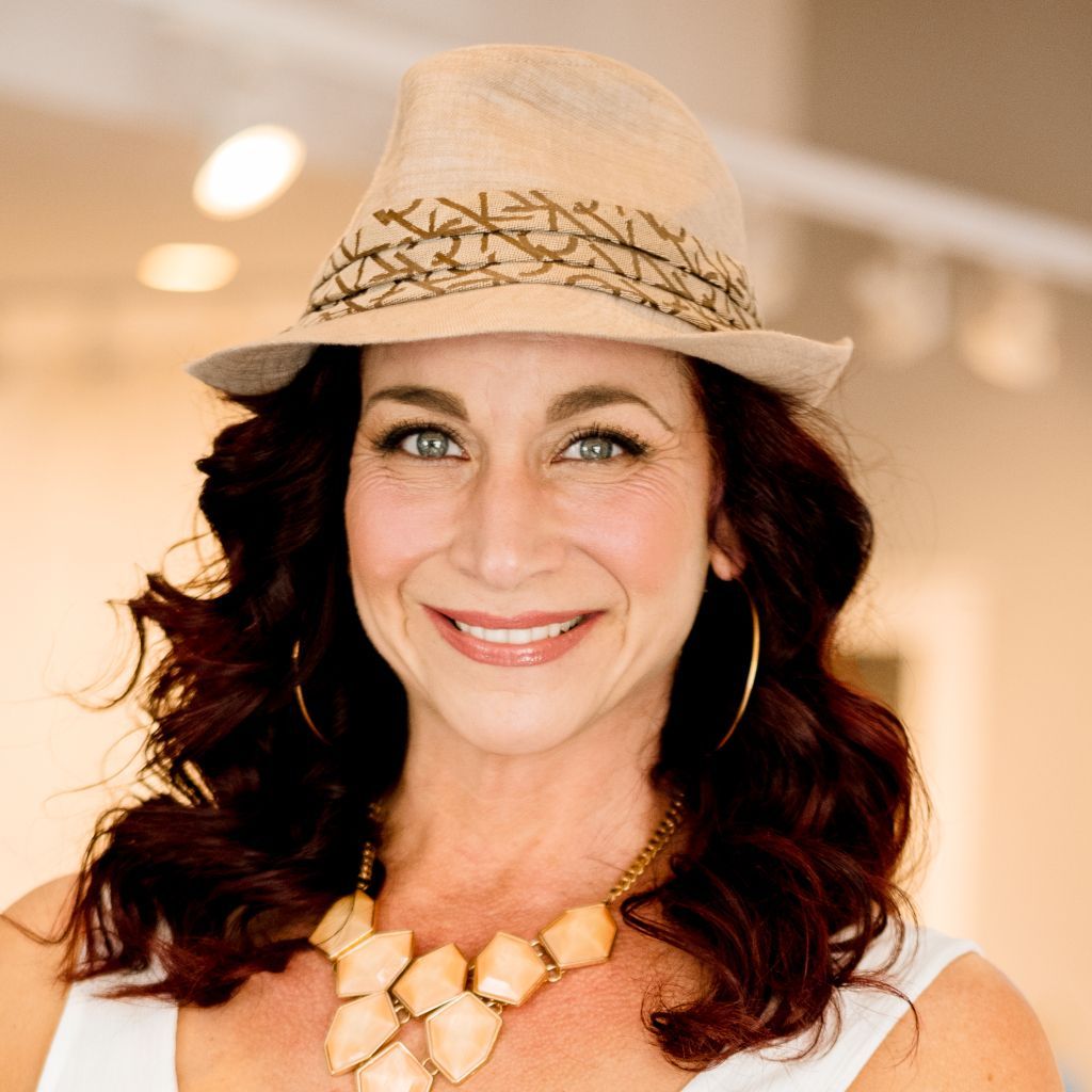 Smiling woman in a beige hat, gold necklace, and white top against a warm indoor background