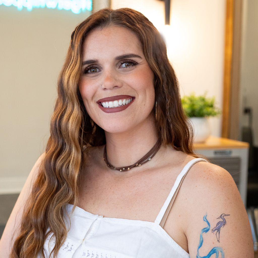 Smiling woman in a white top with a blue floral tattoo, standing indoors near a plant.