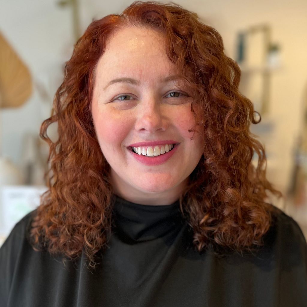 Smiling woman with curly red hair in a black top against a softly blurred indoor background
