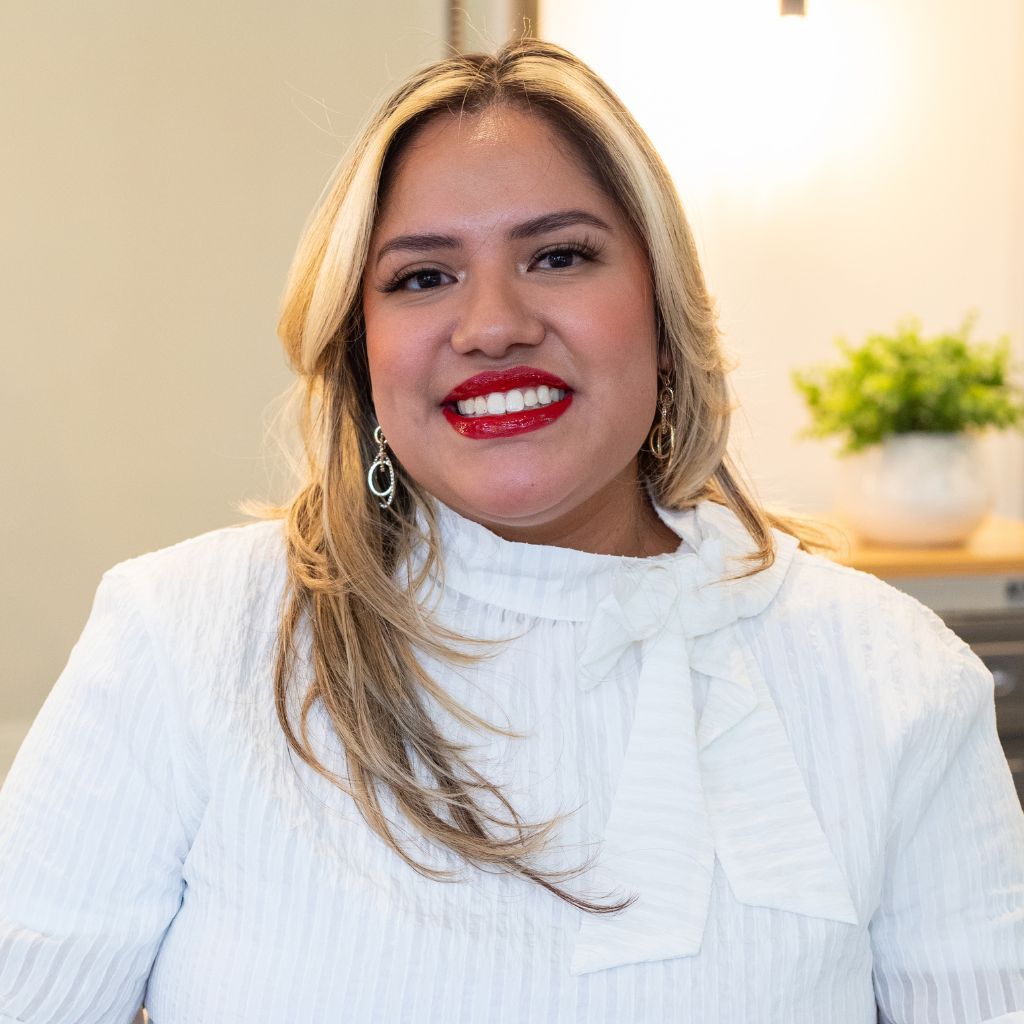 Smiling woman in a white blouse with blonde hair and red lipstick, standing indoors.
