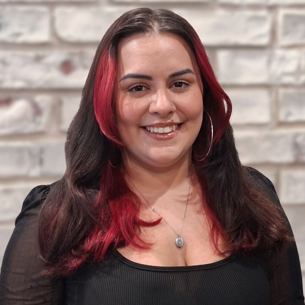 Smiling woman with dark hair and red highlights, wearing a black top against a brick wall background