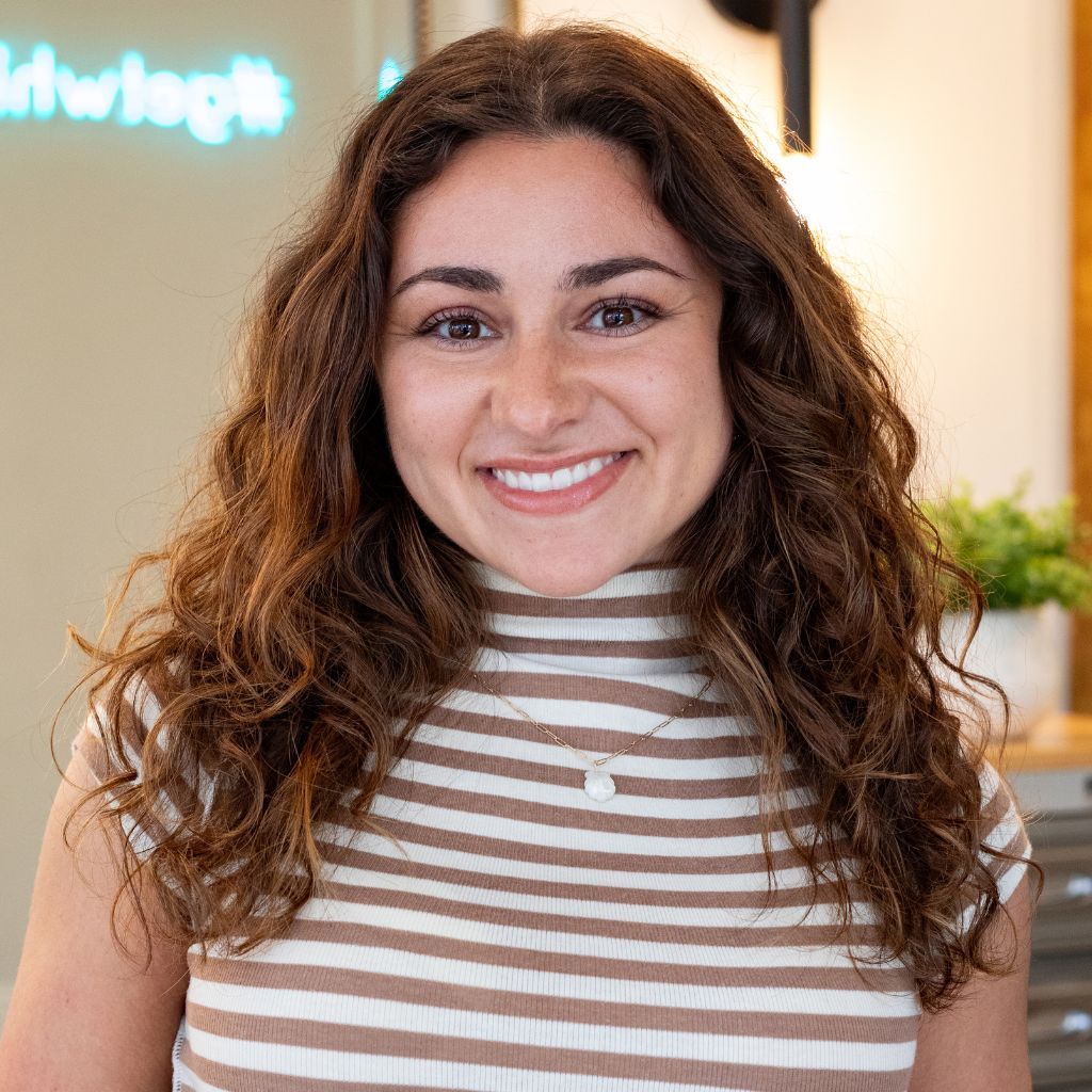 Smiling woman with curly hair in a striped sleeveless top, standing indoors in a bright office.