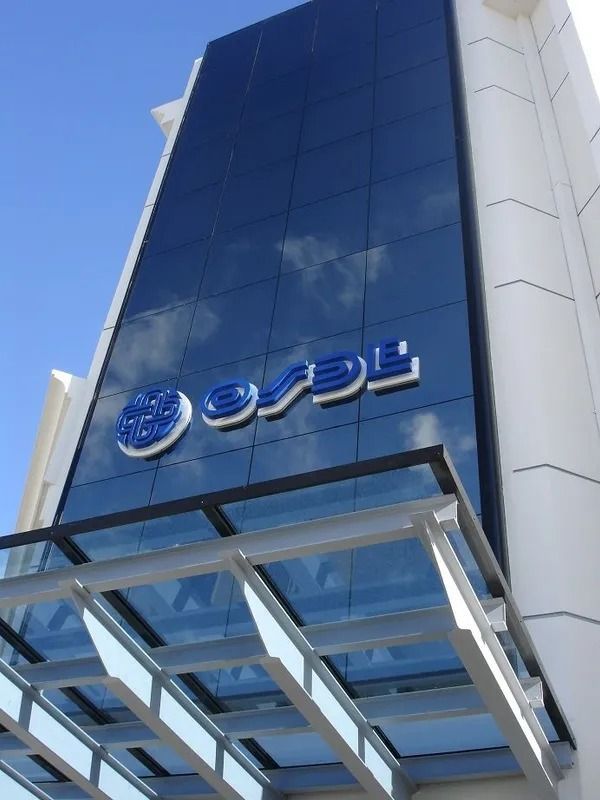 Fachada de un edificio de oficinas de cristal azul con letreros en árabe y una marquesina de cristal contra el cielo.