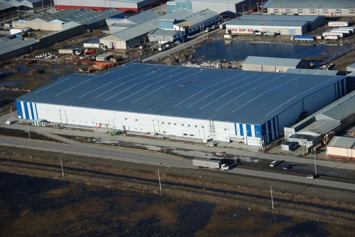 Vista aérea de un gran almacén blanco con techo azul en una zona industrial cerca de una carretera.