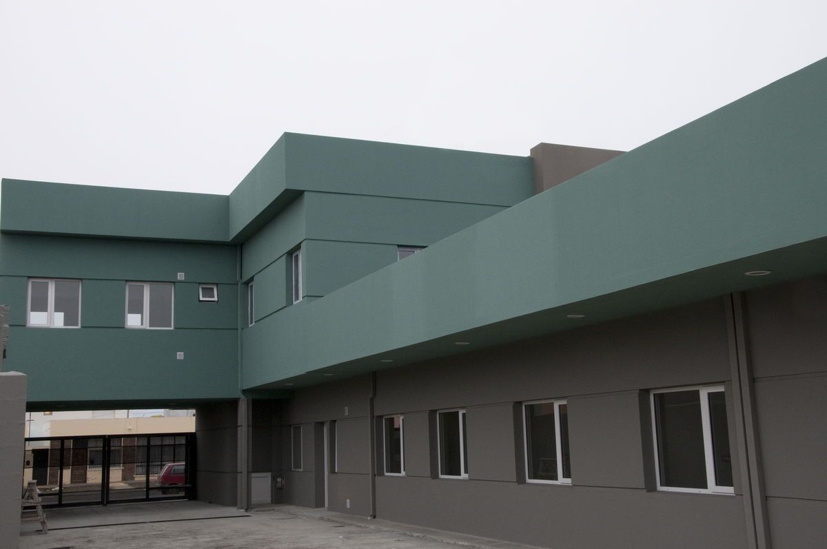 Exterior de edificio verde y gris con ventanas y una pasarela cubierta en un día nublado.