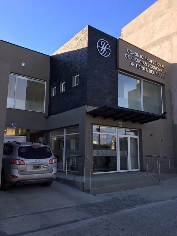 Entrada de un edificio moderno de color gris oscuro con puertas de cristal, un letrero encima y un coche plateado aparcado bajo un cielo azul.