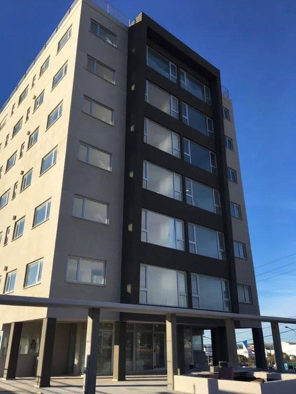Edificio de apartamentos moderno de varias plantas con fachada beige y gris oscuro bajo un cielo azul claro.