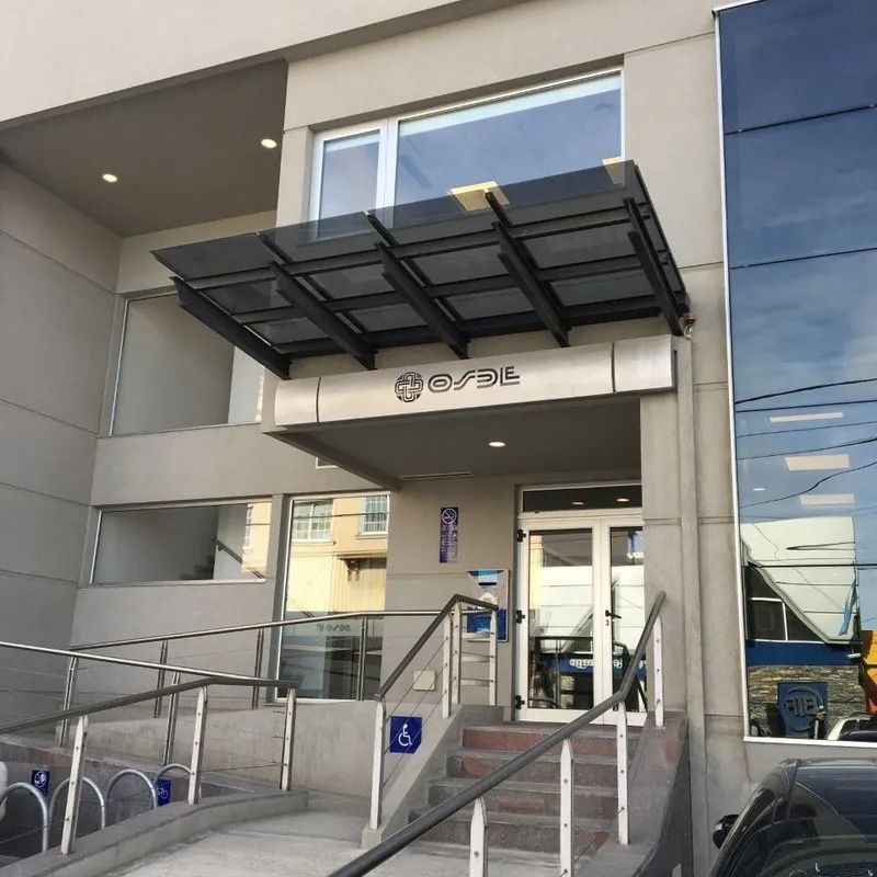 Entrada del edificio con toldo de cristal, rampa, escaleras y señal de accesibilidad cerca de la puerta.