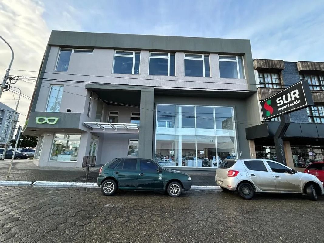 Edificio moderno de color gris con grandes escaparates de cristal y dos coches aparcados en una calle empedrada mojada.