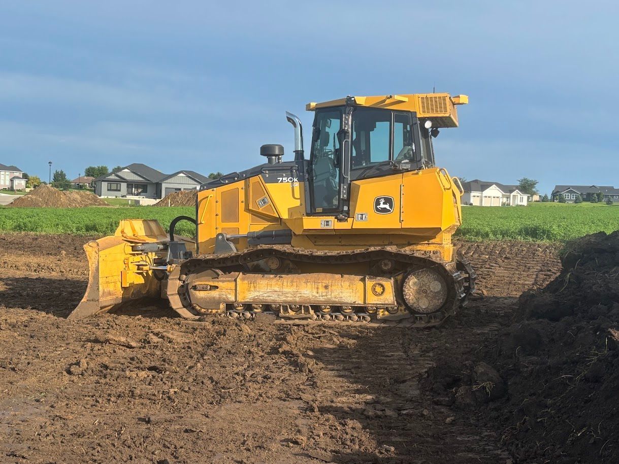 Yellow bulldozer clearing muddy field under a blue sky, with houses in the background