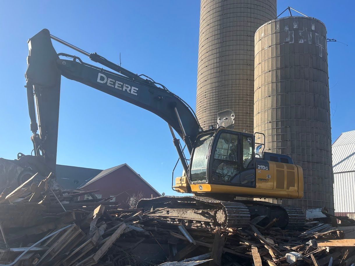 Yellow Deere excavator demolishing rubble beside large concrete silos under a clear blue sky