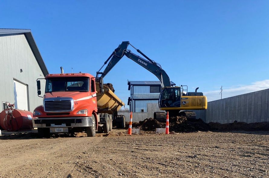 Orange dump truck and yellow excavator working on a gravel lot beside industrial buildings.