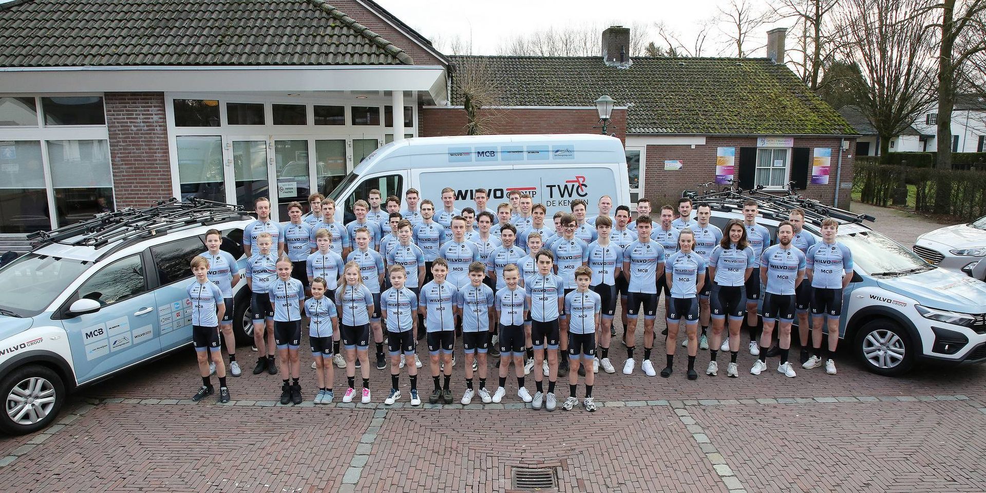 Een groep mensen in lichtblauwe uniformen poseert voor voertuigen en gebouwen.