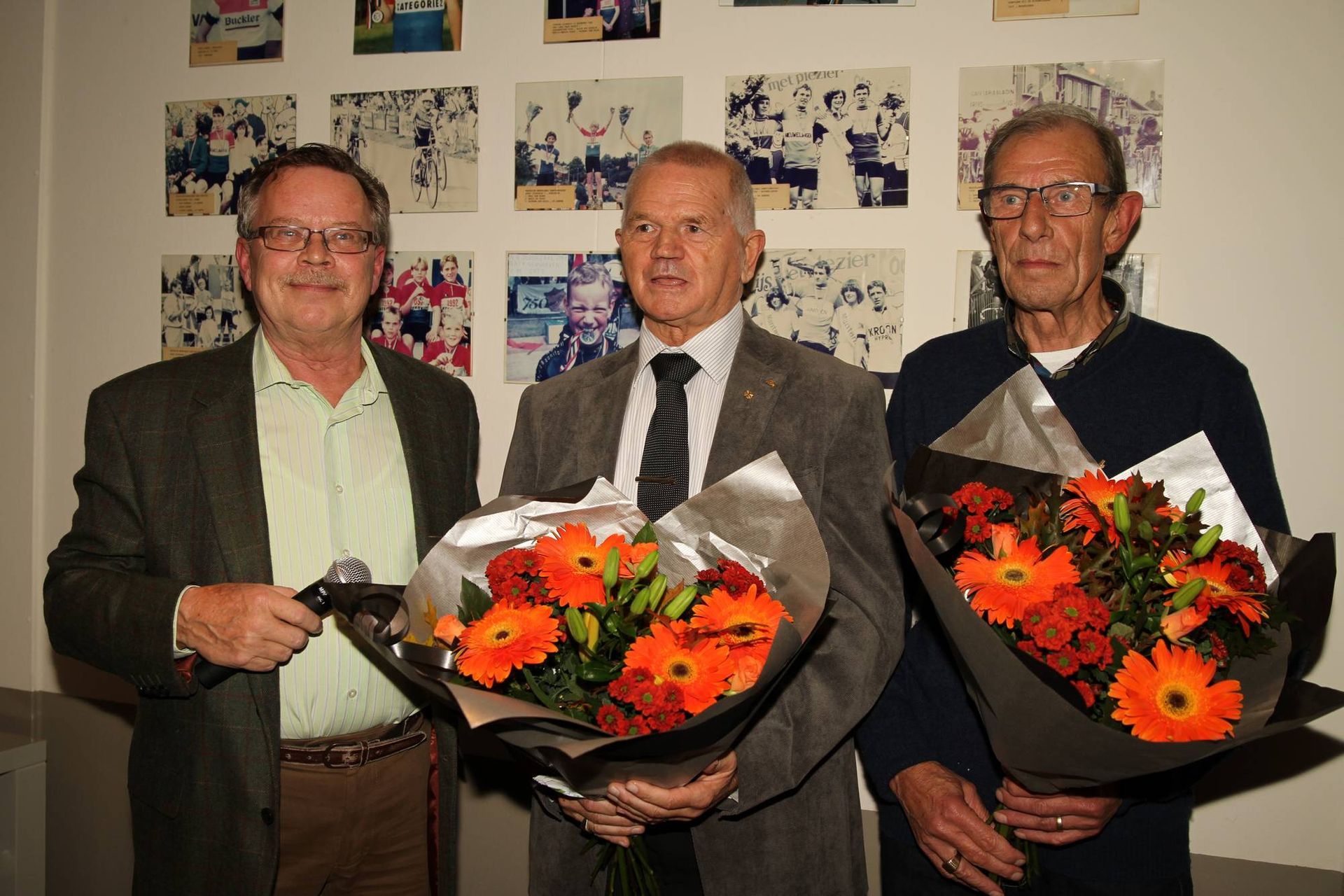 Drie mannen met boeketten staan lachend voor ingelijste foto's.