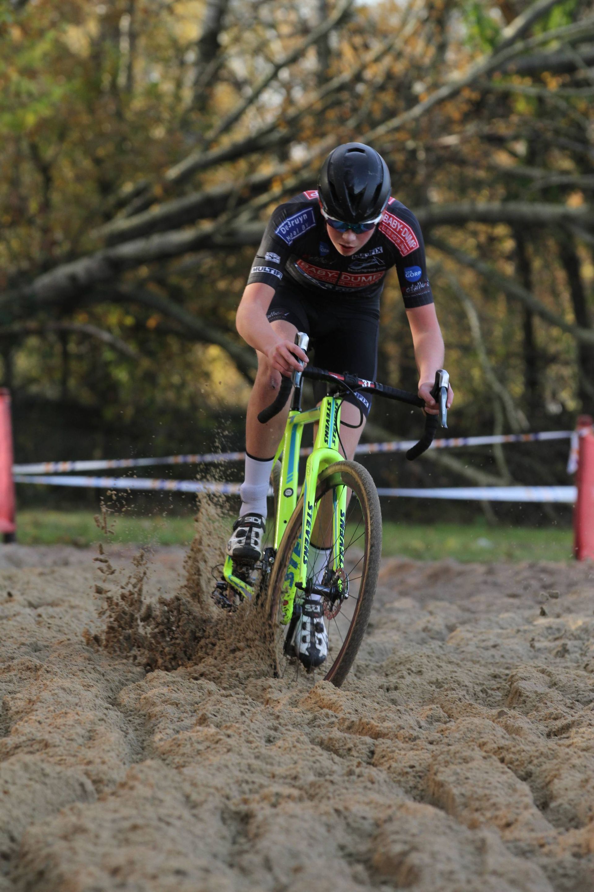 Een cyclocrosser rijdt door de modder, waarbij het voorwiel opspat, en draagt een zwarte helm en een zwart shirt.