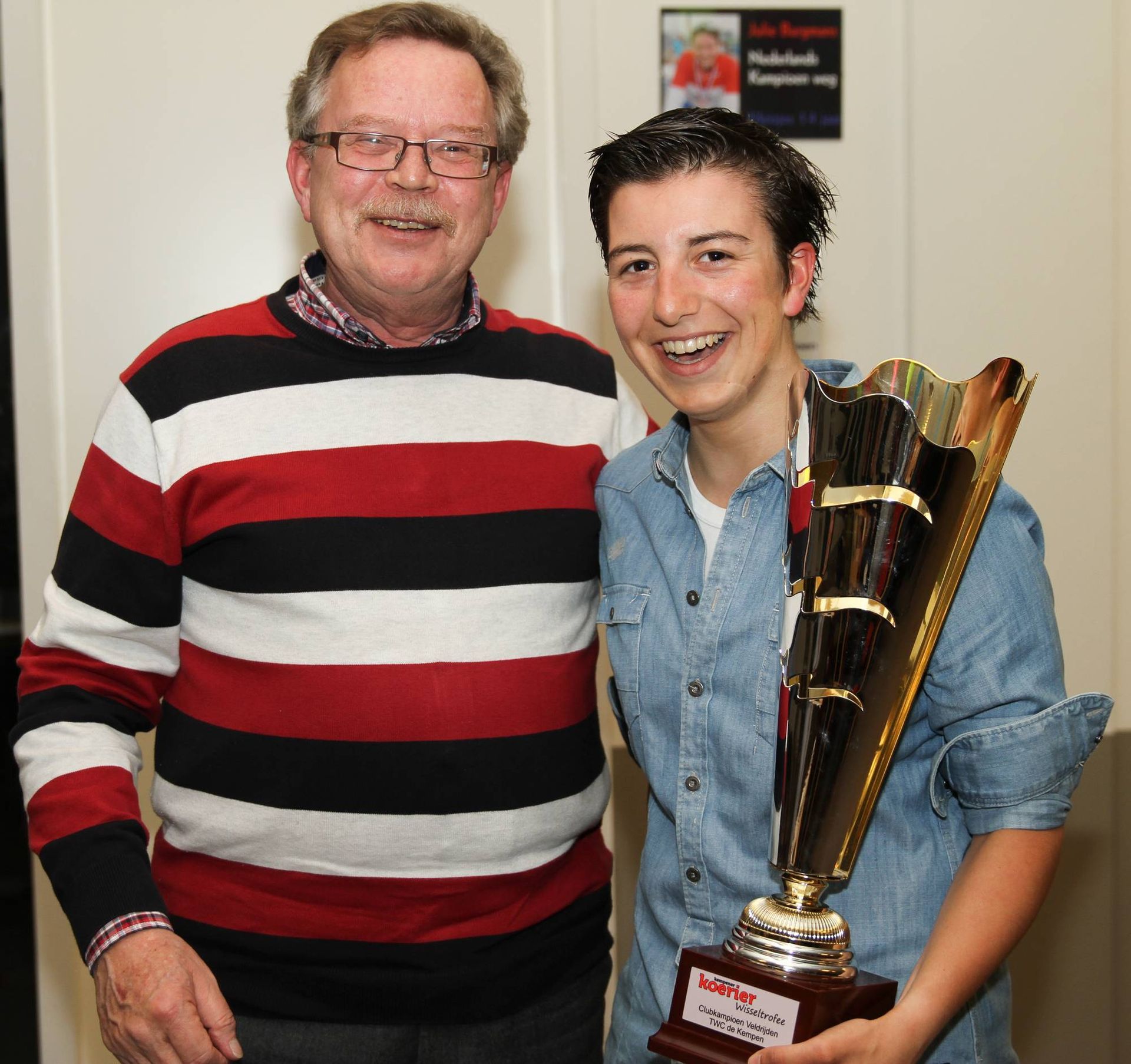 Een man en een jongere glimlachen en houden een trofee vast. De man draagt een bril en een rood-zwarte trui.