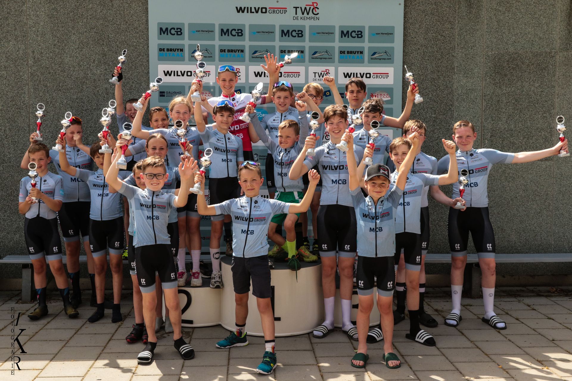 Vier wielrenners staan ​​op een podium met boeketten in hun handen. Een vrouw met een sjerp staat bij hen. 