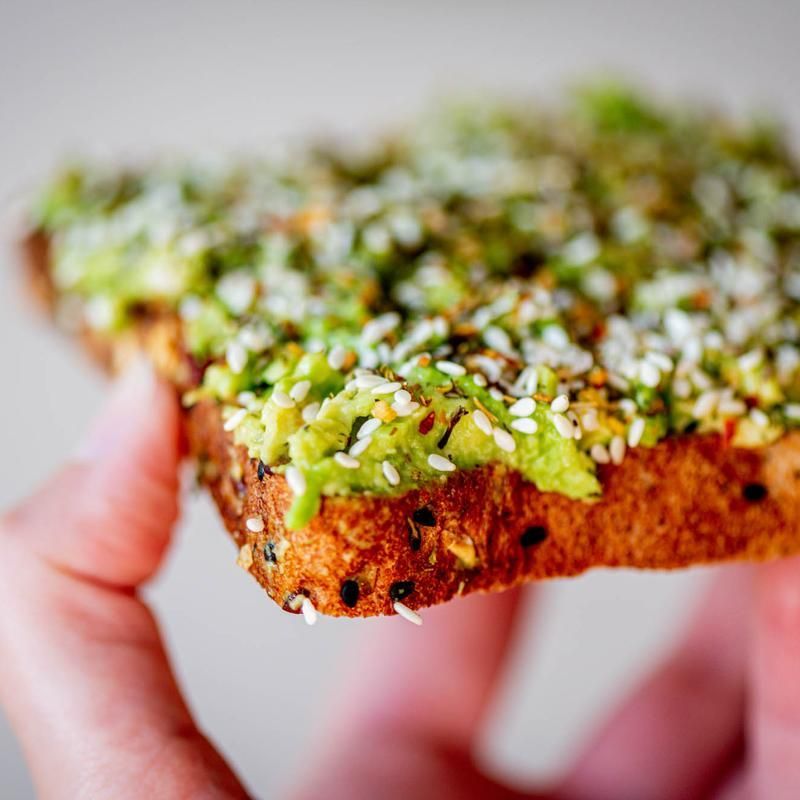Hand holding a slice of toast topped with mashed avocado, seeds, and herbs