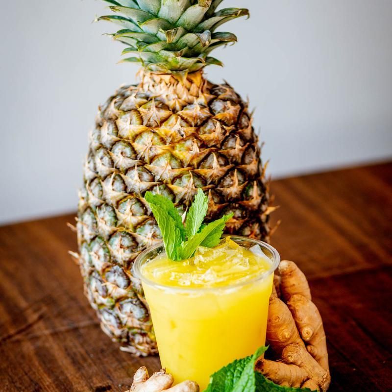 Pineapple with a yellow smoothie topped with mint, beside a piece of ginger on a wooden table.
