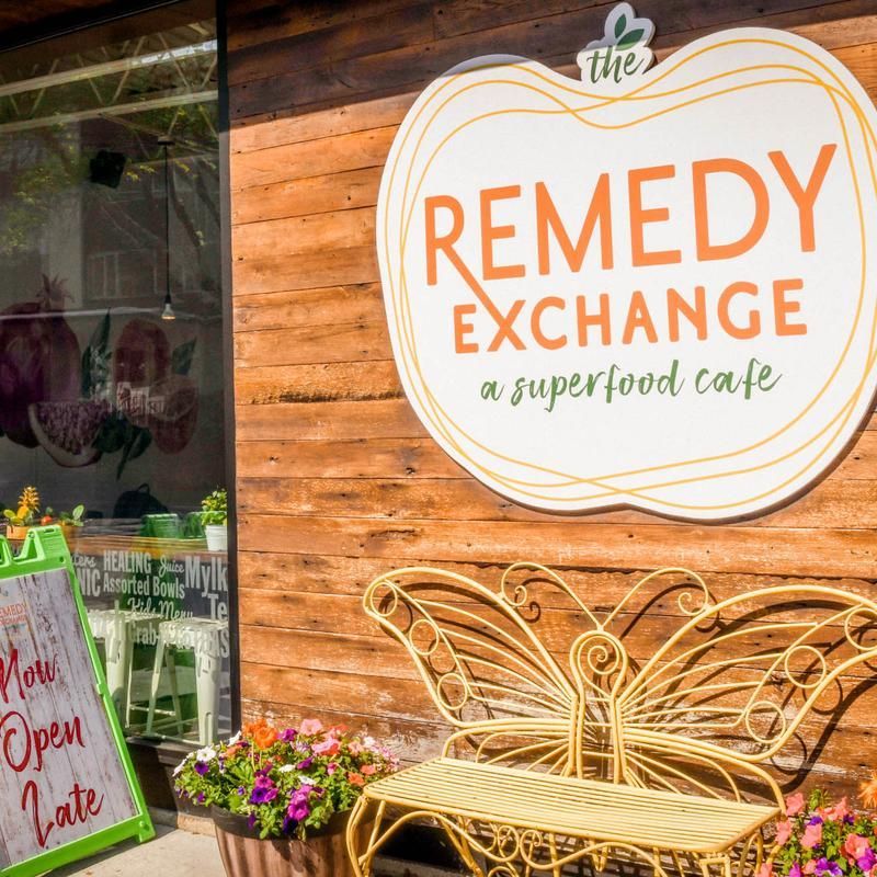 Exterior sign for Remedy Exchange superfood cafe on a wooden storefront, with a wicker bench and flowers.