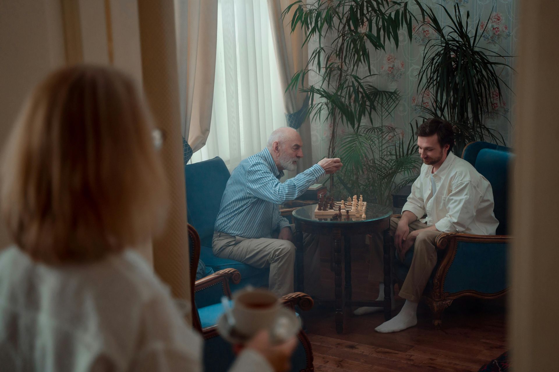Older man playing chess with a younger man, looking on. Seen through a mirror; person in foreground.