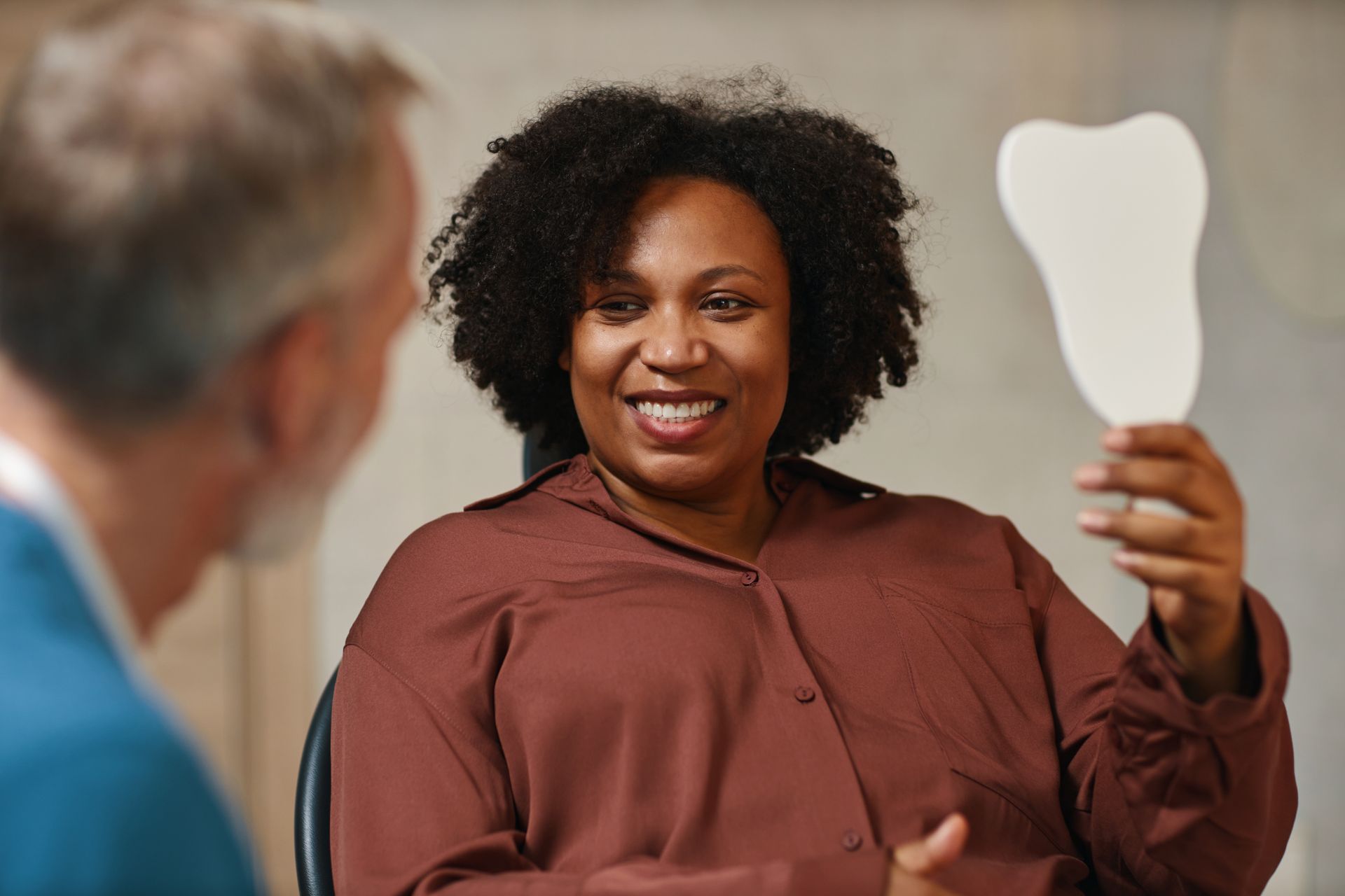 A smiling person in a brown shirt holds a white mirror to inspect their teeth while sitting with a professional.