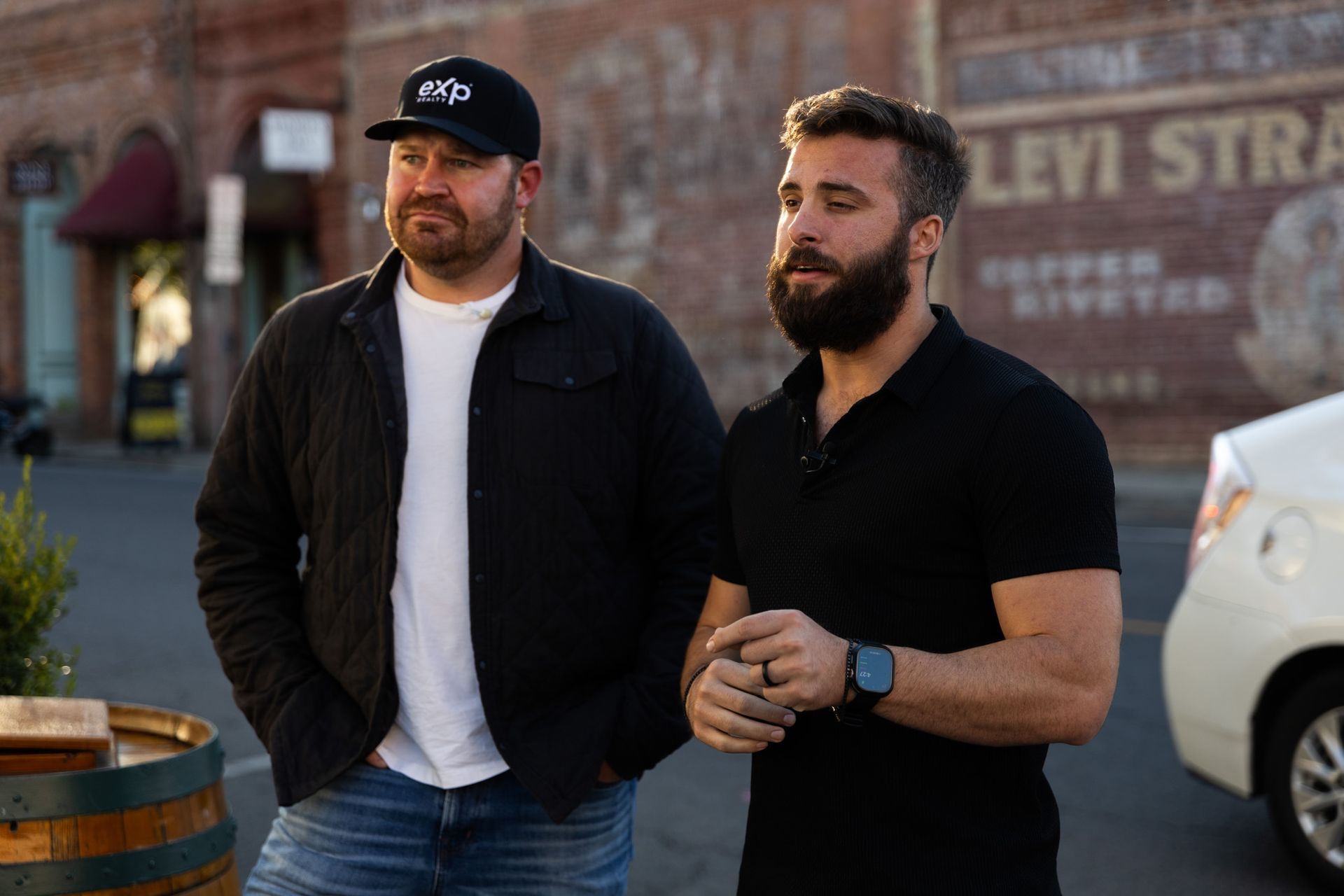 Two men are standing next to each other on a street in front of a building.