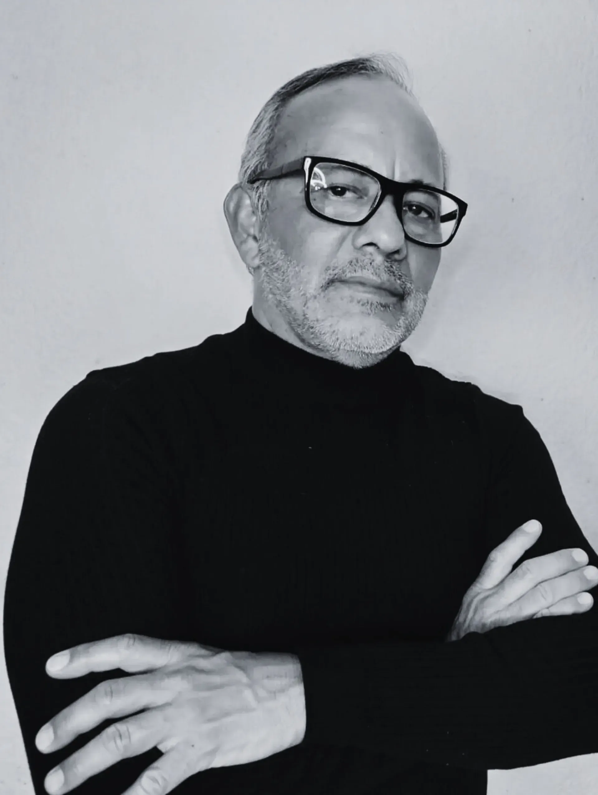 Un hombre con gafas y barba aparece de pie con los brazos cruzados en una fotografía en blanco y negro.