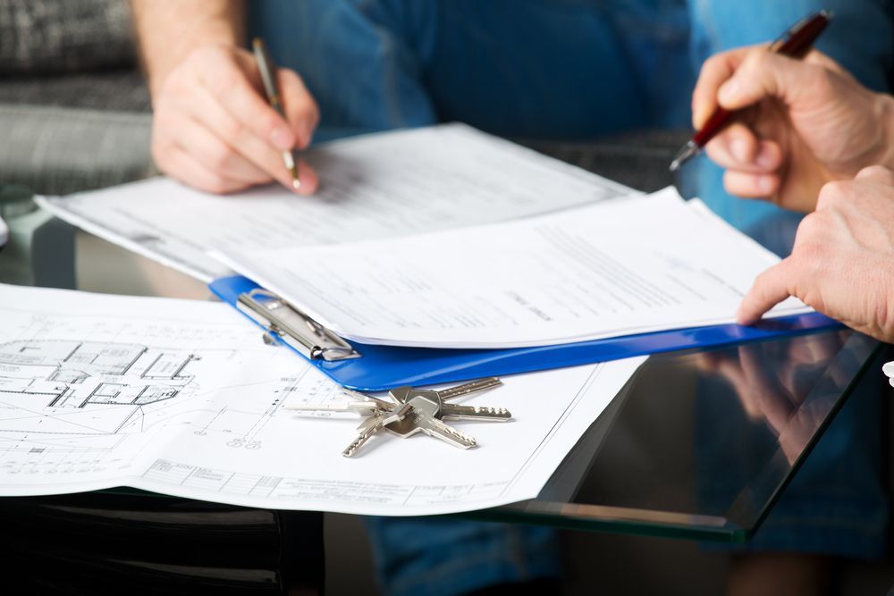 Two People Signing Paperwork With Keys and Blueprints on a Table — Property Transfer Professionals In Wyong, NSW