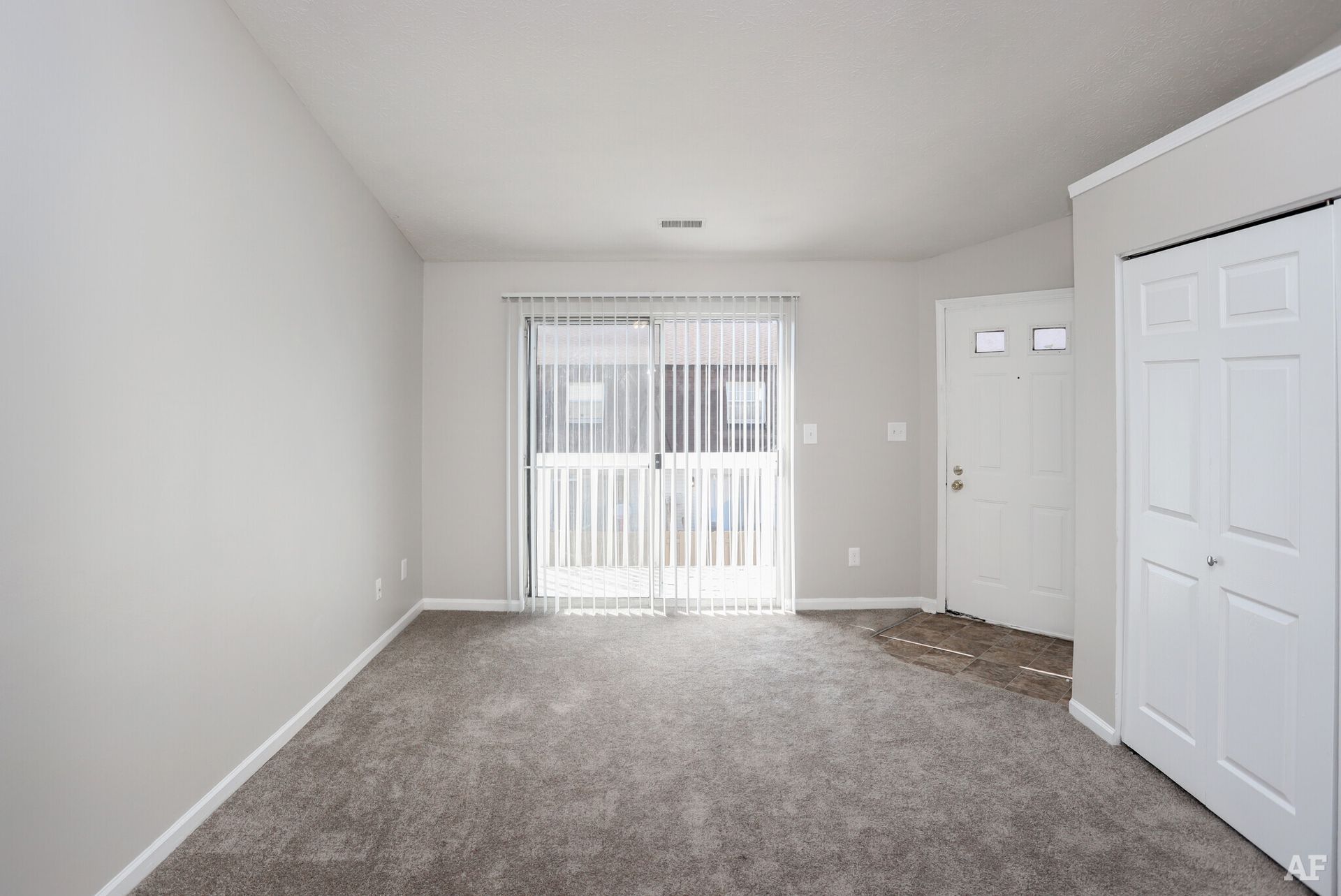 Empty living room with grey carpet, sliding glass door, white door, and closet.