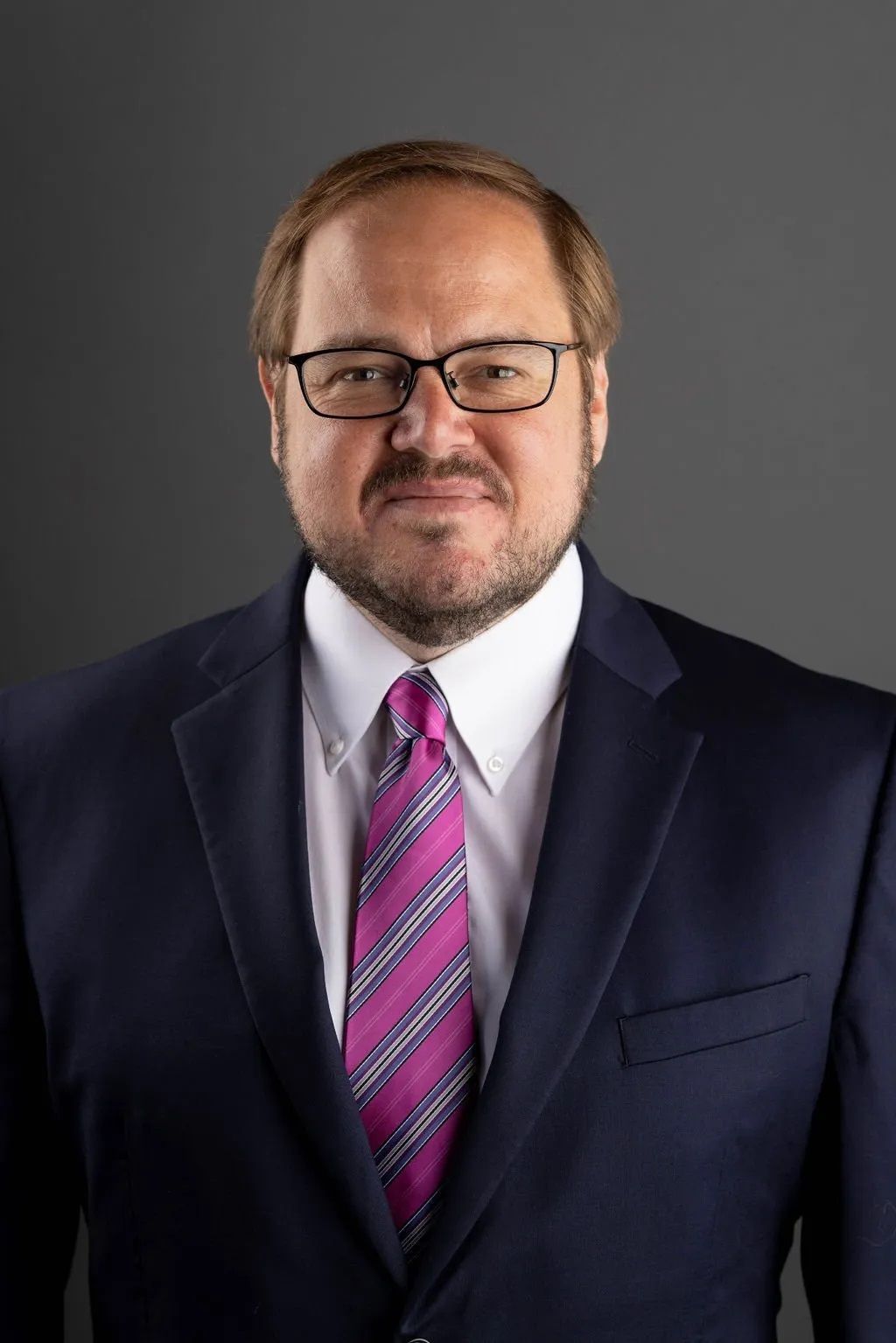Portrait of a man in a dark suit, white shirt, and pink striped tie against a gray background