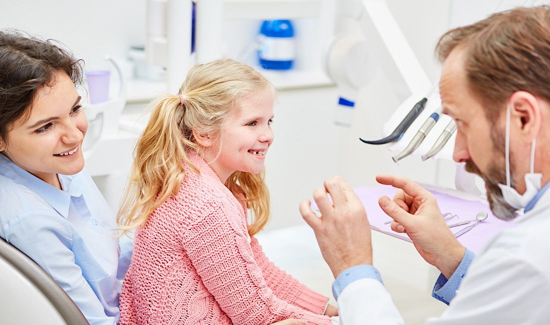 Little girl sitting on mom's lap at the dentist, smiling at dentist while dentist teaches little girl all about teeth.