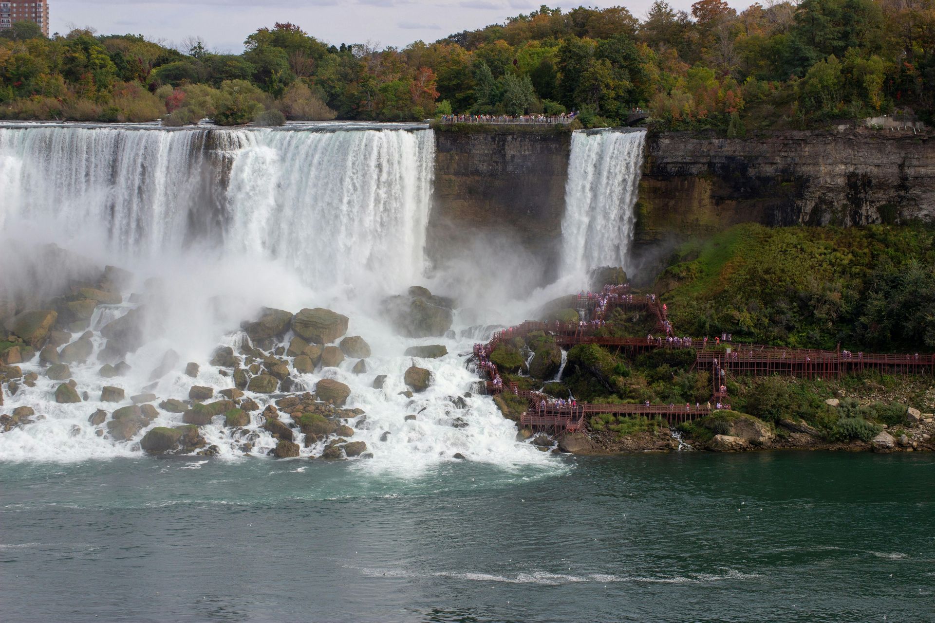 Wide waterfall cascading over a rocky cliff into a green river, with autumn trees above.