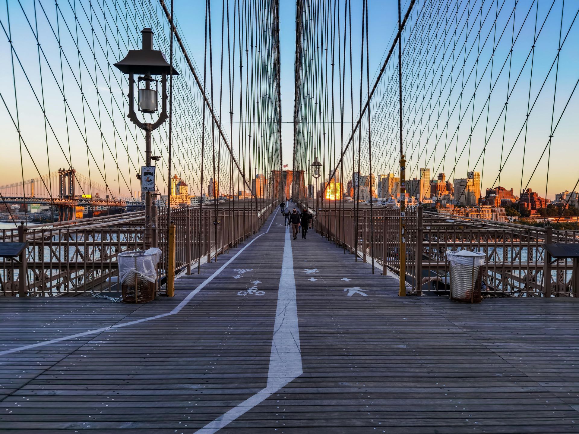 Brooklyn Bridge walkway at sunset, with cables framing the path and city lights in the distance