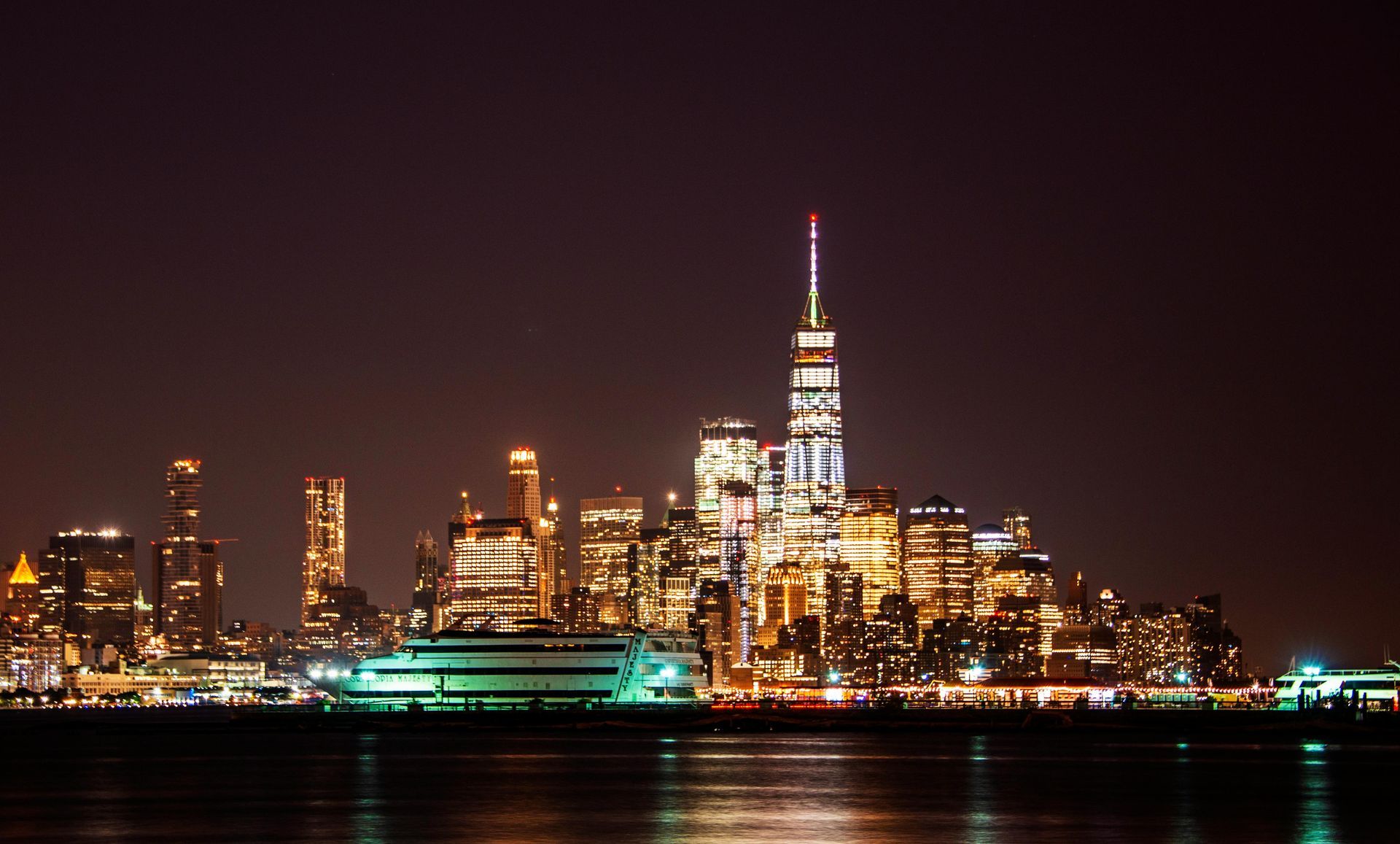 Nighttime city skyline with lit skyscrapers reflecting on water, featuring a tall central tower.
