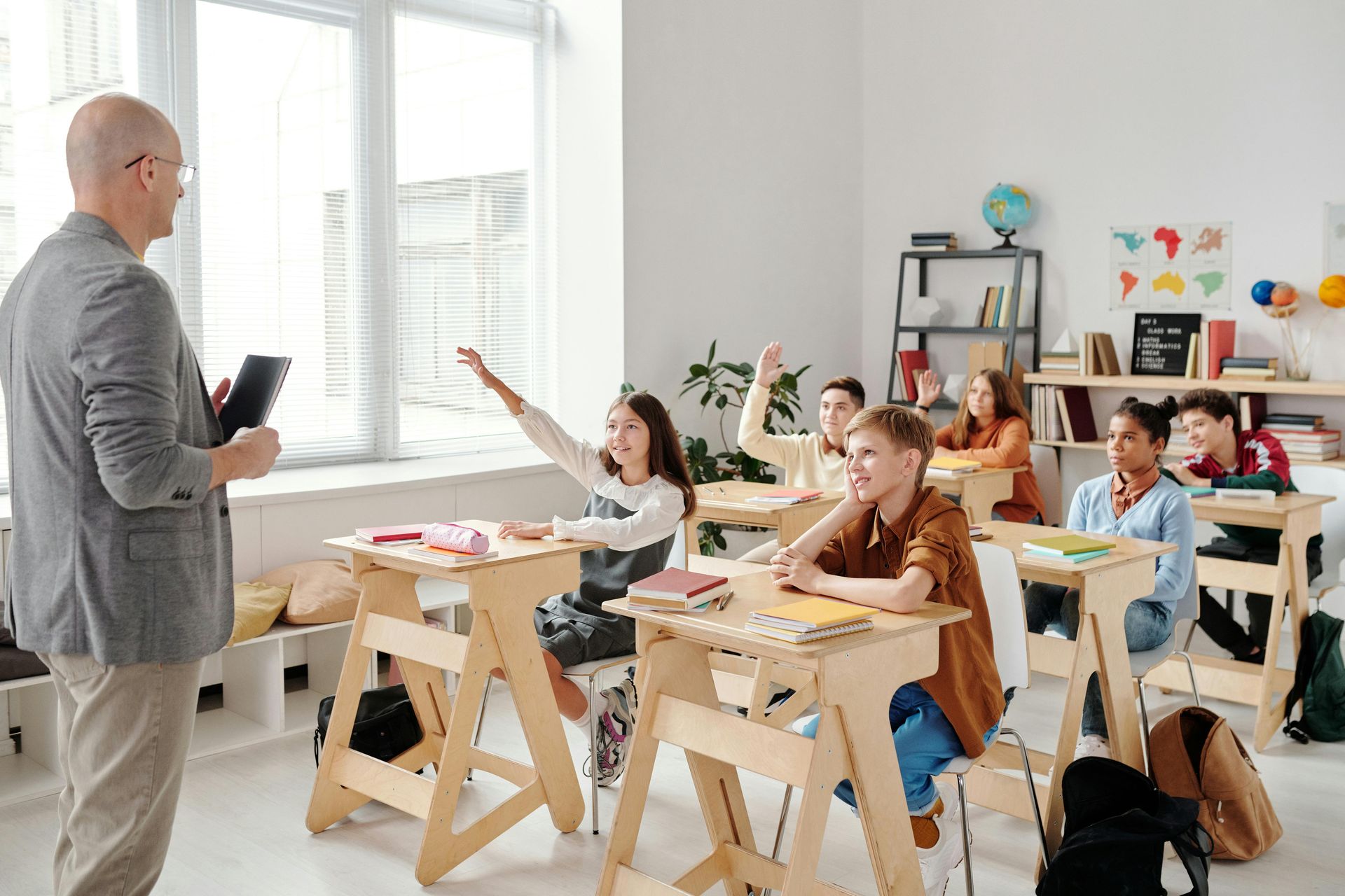 A teacher stands facing a group of students in a classroom with wooden desks, some of whom have their hands raised.