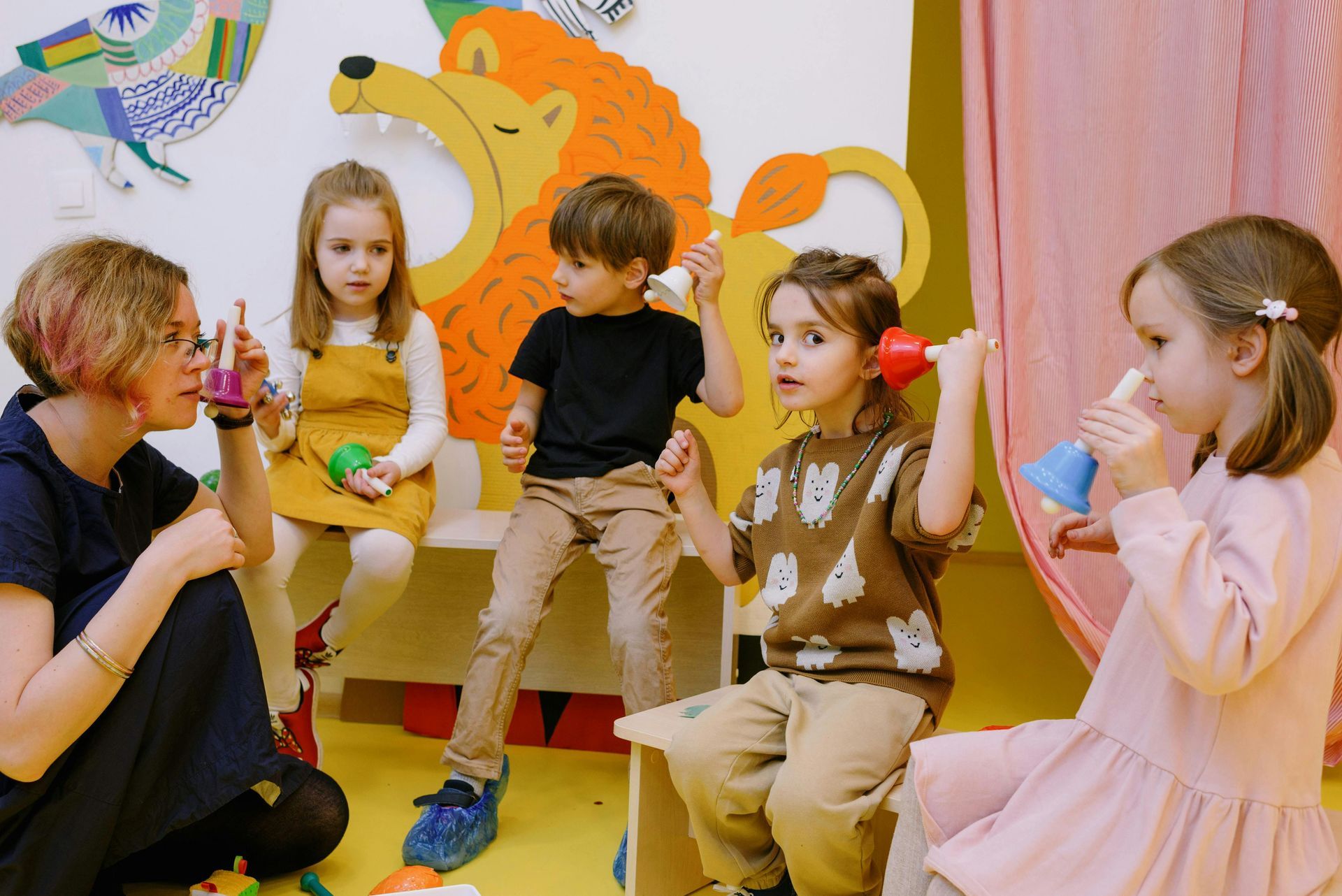 A teacher and four children sit in a brightly decorated room, playing musical bells held to their ears.