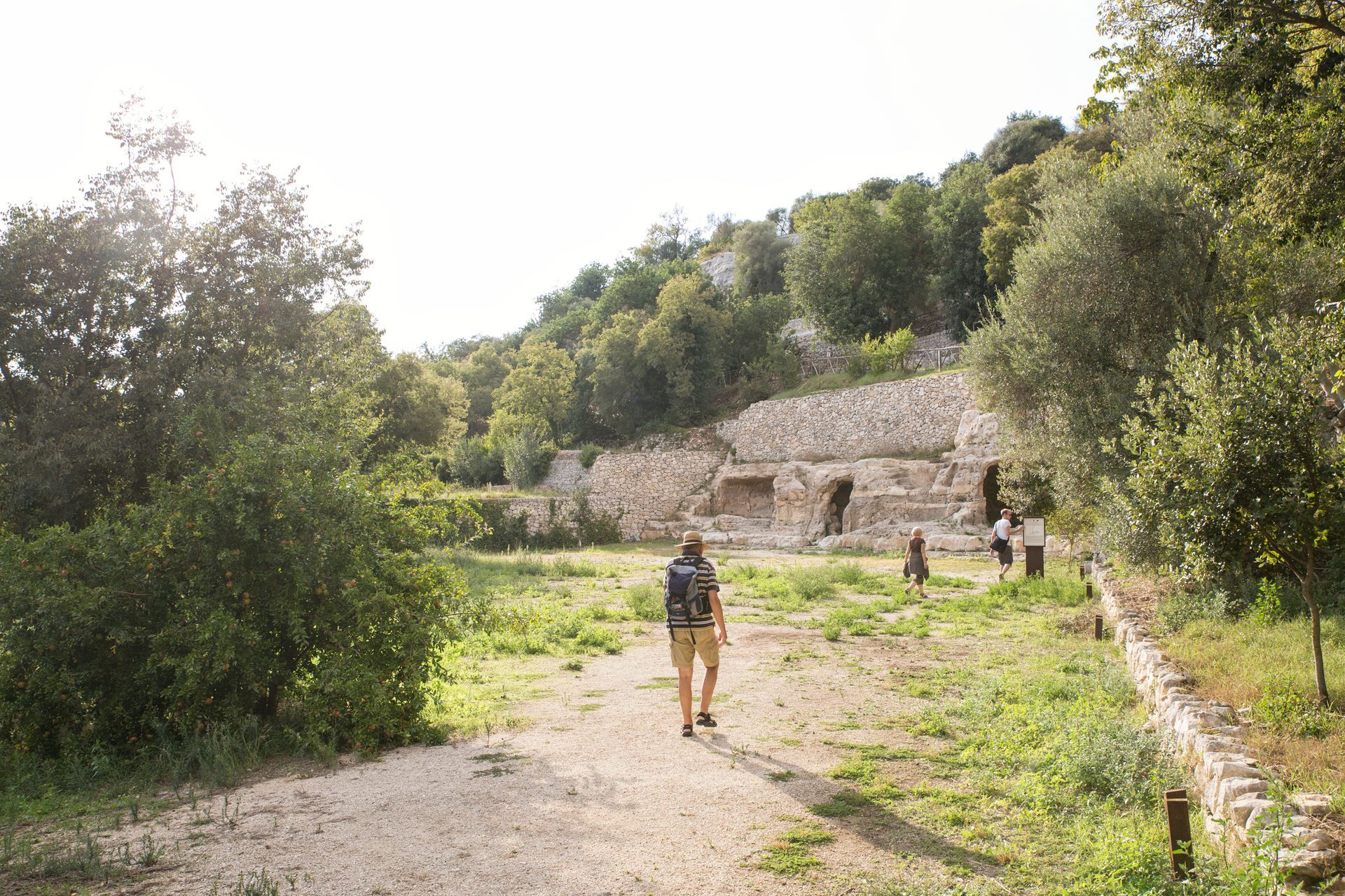 Uomo che cammina verso rovine di pietra, sentiero erboso, alberi, luce del sole.