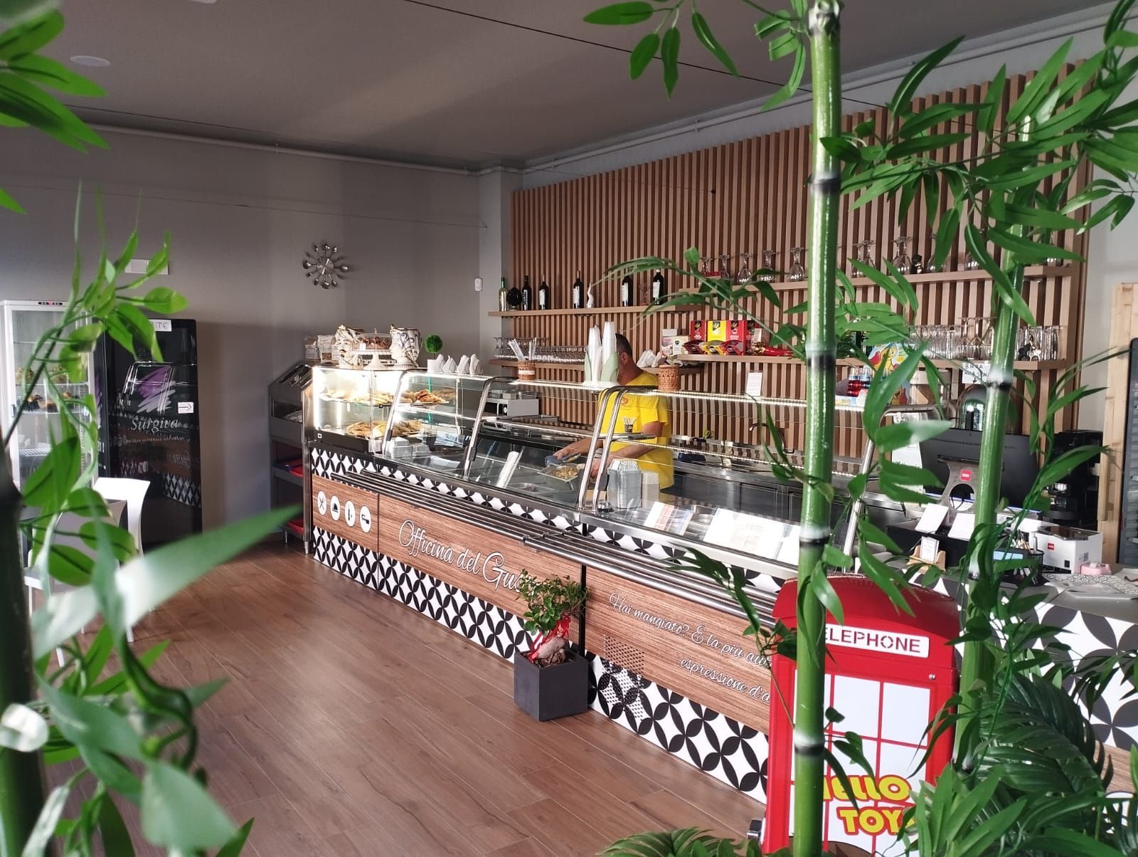Interno del bar: bancone in legno con cibo, piante di bambù, facciata piastrellata in bianco e nero, scaffali con merce.