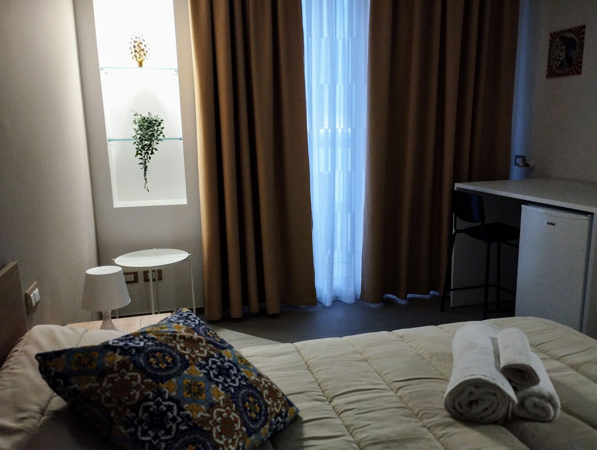 Camera da letto con letto, comodino, finestra con tende, piccola scrivania.