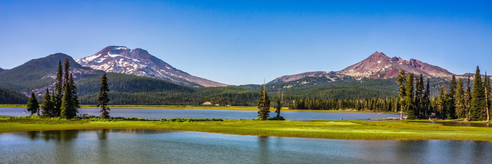 Snowy mountain peaks above a calm lake and green meadow under a clear blue sky