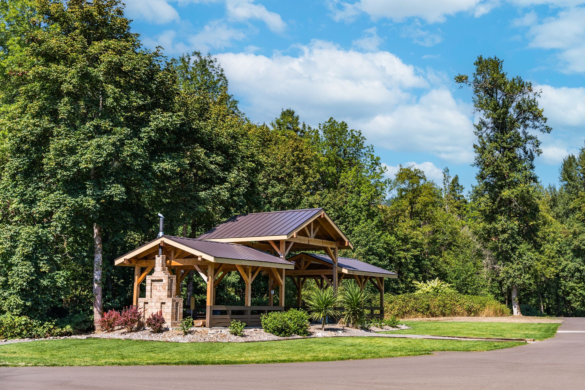 Wooden pavilion with a dark roof in a grassy area, surrounded by trees under a blue sky with clouds.